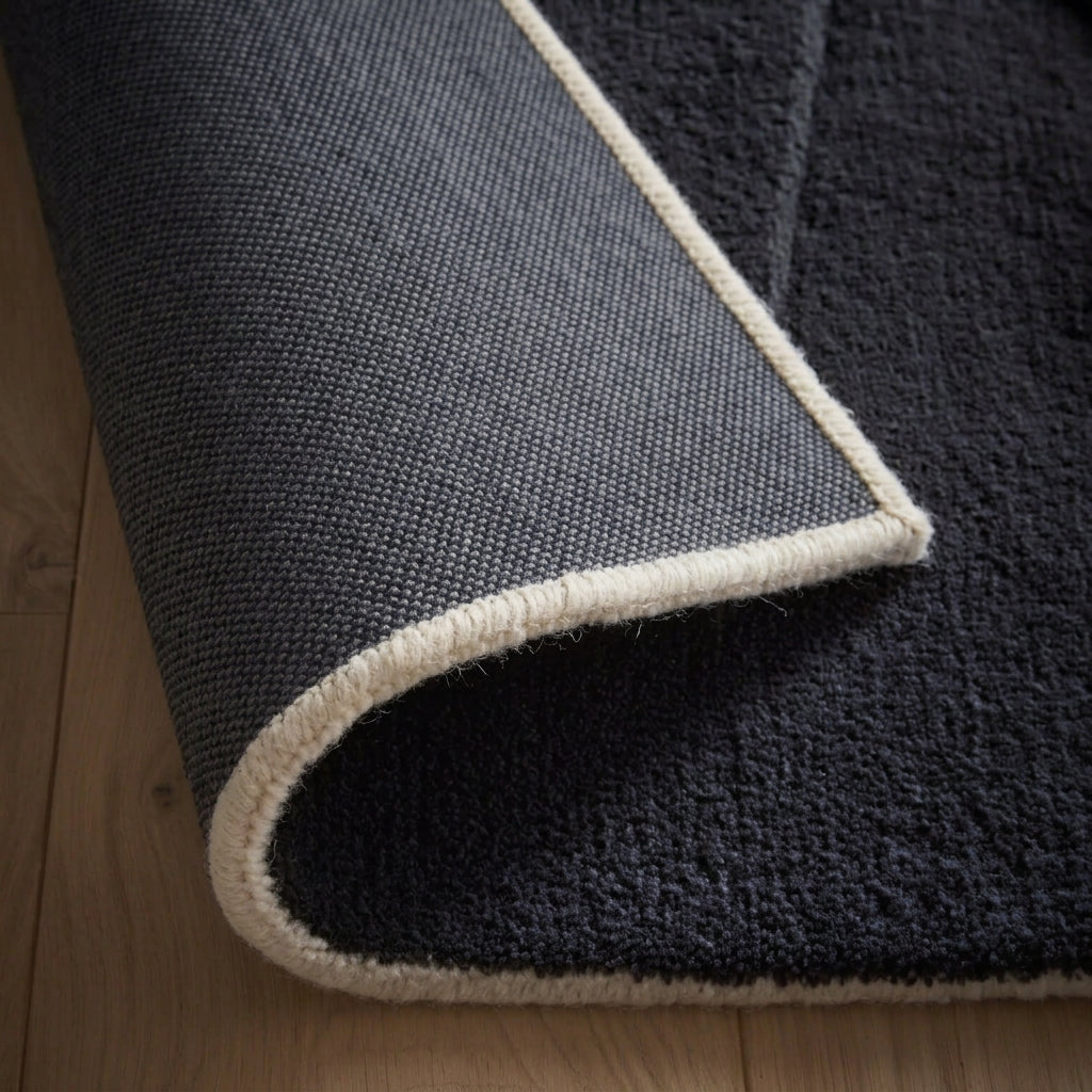A close-up of a deep charcoal black-grey rug on a wood floor, with its corner curled back to reveal its dark charcoal woven backing and a cream whip-stitched edge.