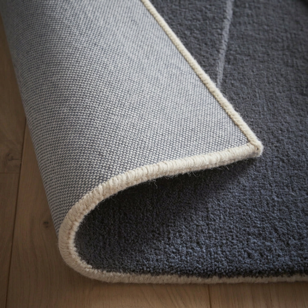 A close-up of a light grey rug on a wood floor, with its corner curled back to reveal its light grey woven backing and a cream whip-stitched edge.